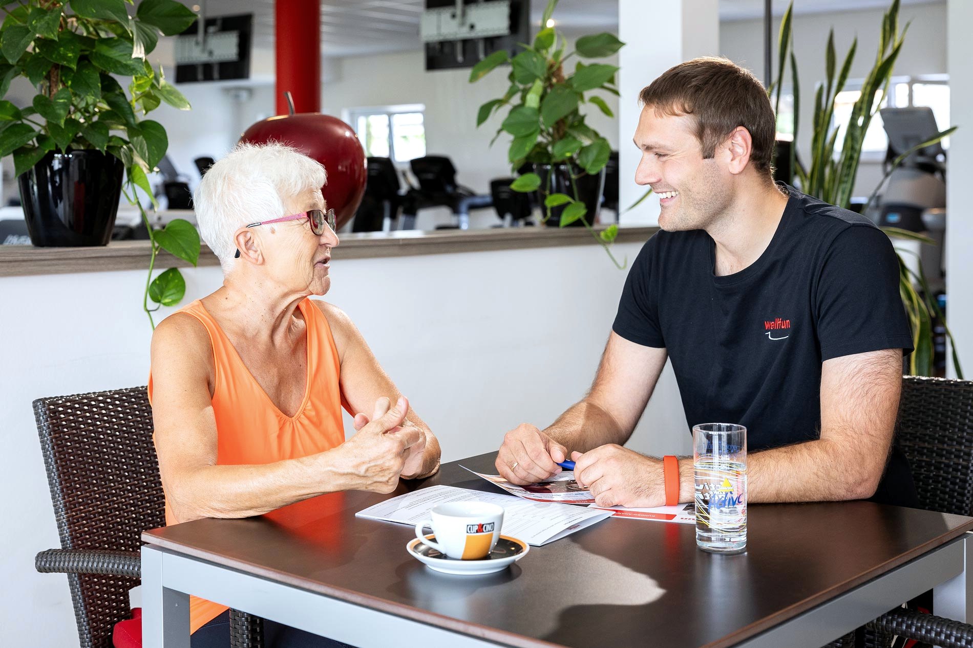 Seniorin im Beratungsgespräch mit einem Trainer, im Hintergrund die Trainingsfläche